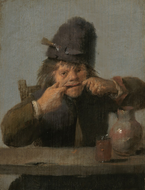 Young man in a hat pulls his mouth wide with fingers at a table.